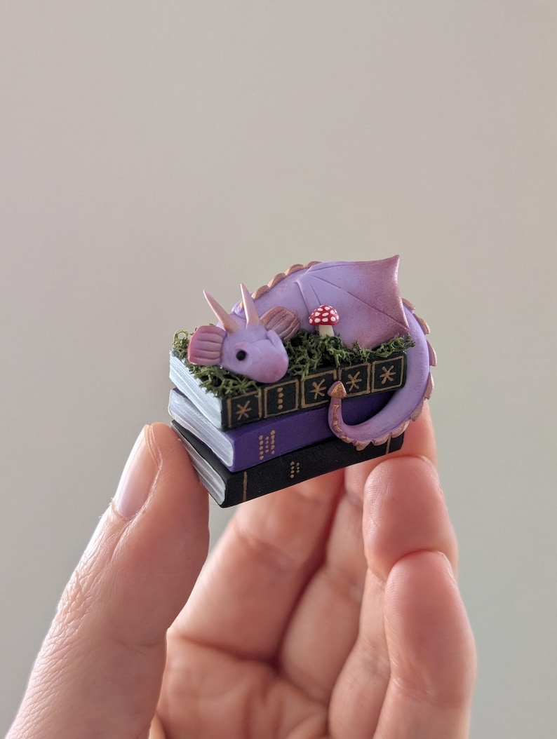 Puede incluir: Una figura de drag&oacute;n p&uacute;rpura en miniatura que descansa sobre una pila de tres libros. El drag&oacute;n tiene una cola larga, alas y cuernos. Una peque&ntilde;a seta roja y blanca est&aacute; cerca. Los libros son negros, morados y blancos.