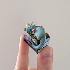 Puede incluir: Una figura de drag&oacute;n azul en miniatura con un hongo con manchas rojas descansa sobre una pila de libros diminutos. El drag&oacute;n est&aacute; enroscado, con sus alas y cola visibles. Los libros son morados y negros, con musgo y hierba verdes alrededor.