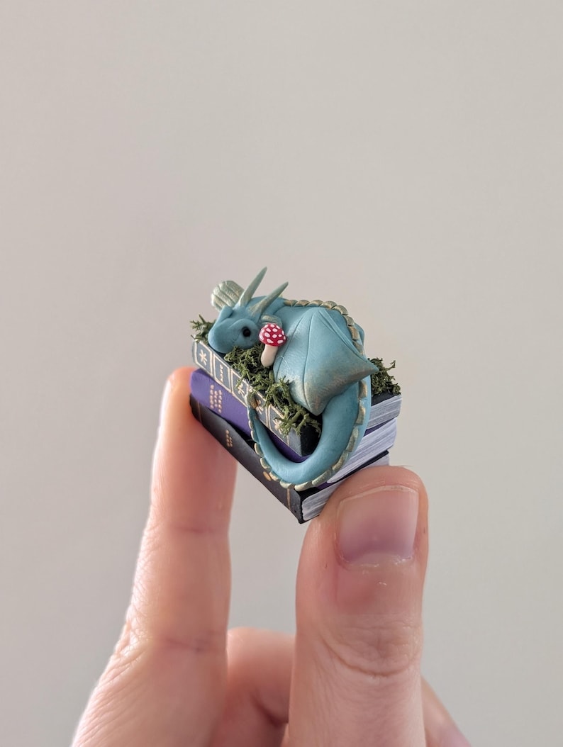 Puede incluir: Una figura de drag&oacute;n azul en miniatura con un hongo con manchas rojas descansa sobre una pila de libros diminutos. El drag&oacute;n est&aacute; enroscado, con sus alas y cola visibles. Los libros son morados y negros, con musgo y hierba verdes alrededor.
