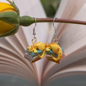 May include: A pair of yellow dragon earrings. Each dragon rests on a stack of miniature books with blue and brown covers. The earrings hang from silver-coloured fish hook ear wires. The background is a blurred open book.
