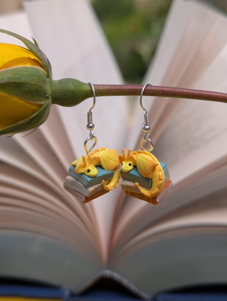 May include: A pair of yellow dragon earrings. Each dragon rests on a stack of miniature books with blue and brown covers. The earrings hang from silver-coloured fish hook ear wires. The background is a blurred open book.