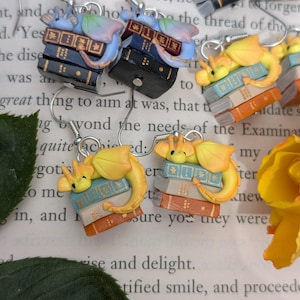 May include: Novelty earrings with a fantasy theme. Each earring features a stack of miniature books with a small dragon figure. The earrings are in various colors, including blue, orange, and black, and have silver hooks. A charming accessory.