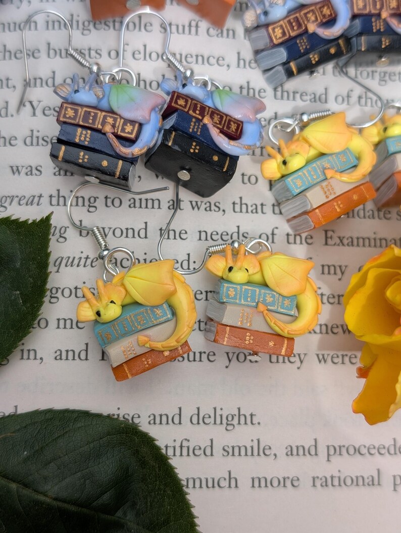 May include: Novelty earrings with a fantasy theme. Each earring features a stack of miniature books with a small dragon figure. The earrings are in various colors, including blue, orange, and black, and have silver hooks. A charming accessory.