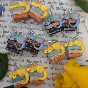 May include: Handmade earrings with a fantasy theme. Each earring features a small yellow dragon resting on a stack of books in various colours. The earrings are suspended from silver-coloured ear wires. The books are in shades of brown, blue, and white.