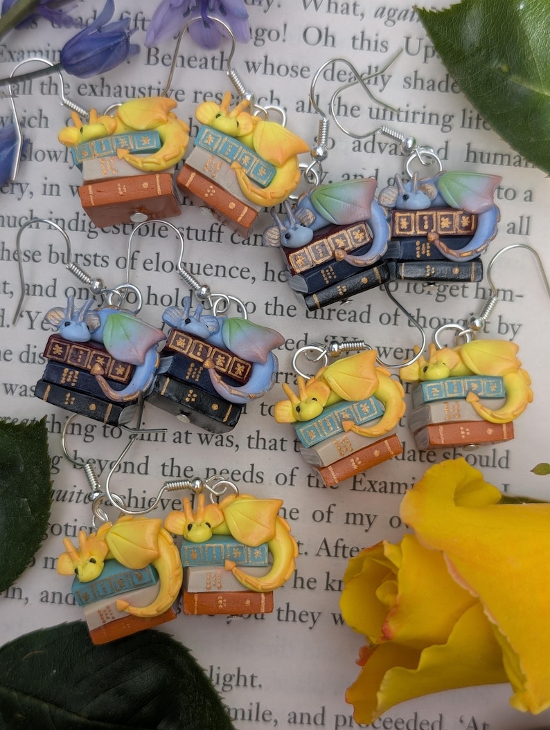 May include: Handmade earrings with a fantasy theme. Each earring features a small yellow dragon resting on a stack of books in various colours. The earrings are suspended from silver-coloured ear wires. The books are in shades of brown, blue, and white.