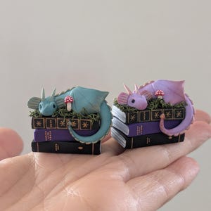 Puede incluir: Dos figuras de dragones en miniatura, una turquesa y otra lavanda, descansan sobre pilas de libros. Cada drag&oacute;n est&aacute; metido entre musgo verde y una seta roja y blanca. Los libros son negros y morados con detalles dorados.