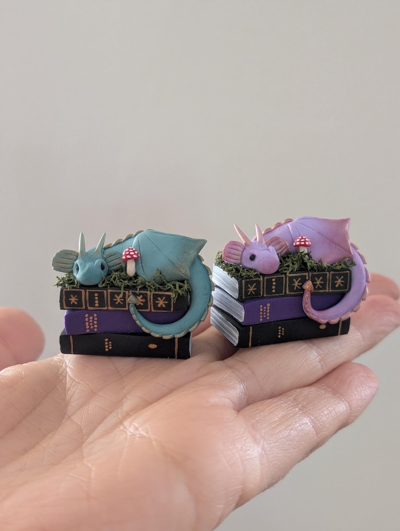 Puede incluir: Dos figuras de dragones en miniatura, una turquesa y otra lavanda, descansan sobre pilas de libros. Cada drag&oacute;n est&aacute; metido entre musgo verde y una seta roja y blanca. Los libros son negros y morados con detalles dorados.