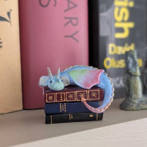 May include: A small, blue and pastel-colored dragon figurine resting on a stack of miniature books. The dragon has a light blue body, with pink and green wings. The books are dark blue, with gold detailing.