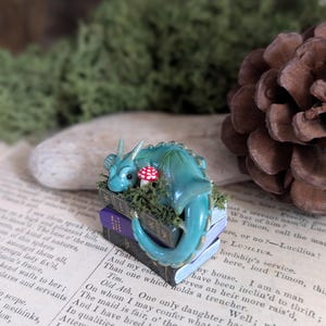 Puede incluir: Figura de drag&oacute;n turquesa sobre una pila de libros en miniatura con una seta roja. El drag&oacute;n tiene una cola enroscada y est&aacute; rodeado de musgo verde. Los libros son negros y morados.