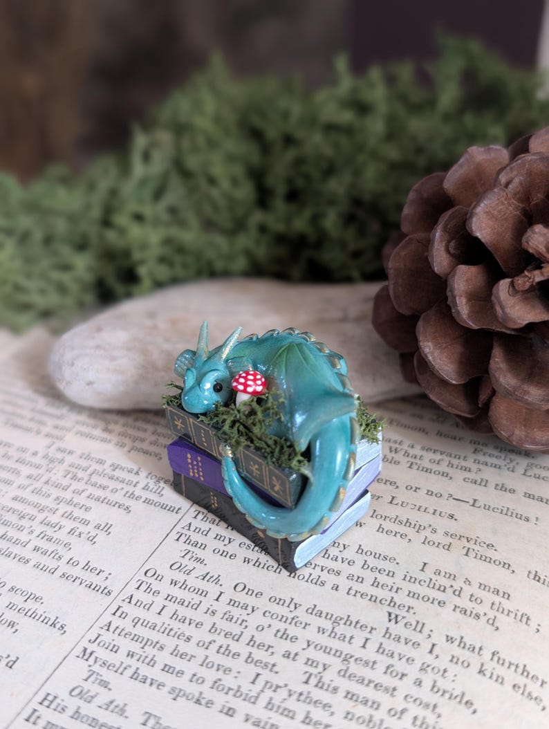 Puede incluir: Figura de drag&oacute;n turquesa sobre una pila de libros en miniatura con una seta roja. El drag&oacute;n tiene una cola enroscada y est&aacute; rodeado de musgo verde. Los libros son negros y morados.
