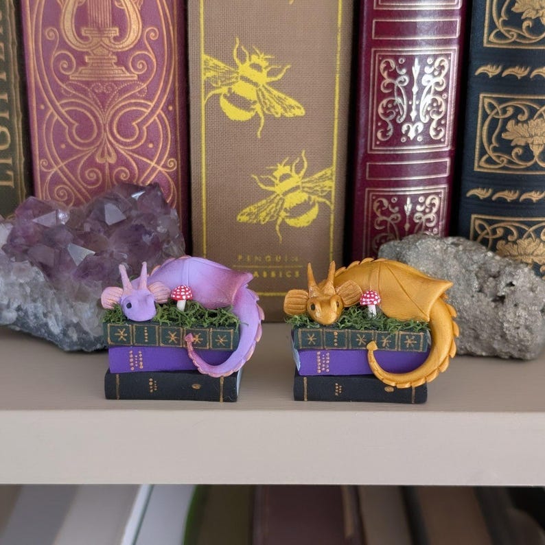 Puede incluir: Dos figuras de dragones en miniatura, una lavanda y otra dorada, descansan sobre pilas de libros morados y negros. Cada drag&oacute;n tiene un peque&ntilde;o hongo y detalles de hierba. El fondo presenta una colecci&oacute;n de libros antiguos.