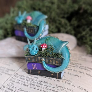Puede incluir: Dos esculturas de fantas&iacute;a en miniatura. Cada una presenta un drag&oacute;n verde azulado enroscado alrededor de una pila de libros. Los libros son negros y morados, con un peque&ntilde;o hongo y vegetaci&oacute;n encima. El drag&oacute;n tiene una cara y una cola detalladas.
