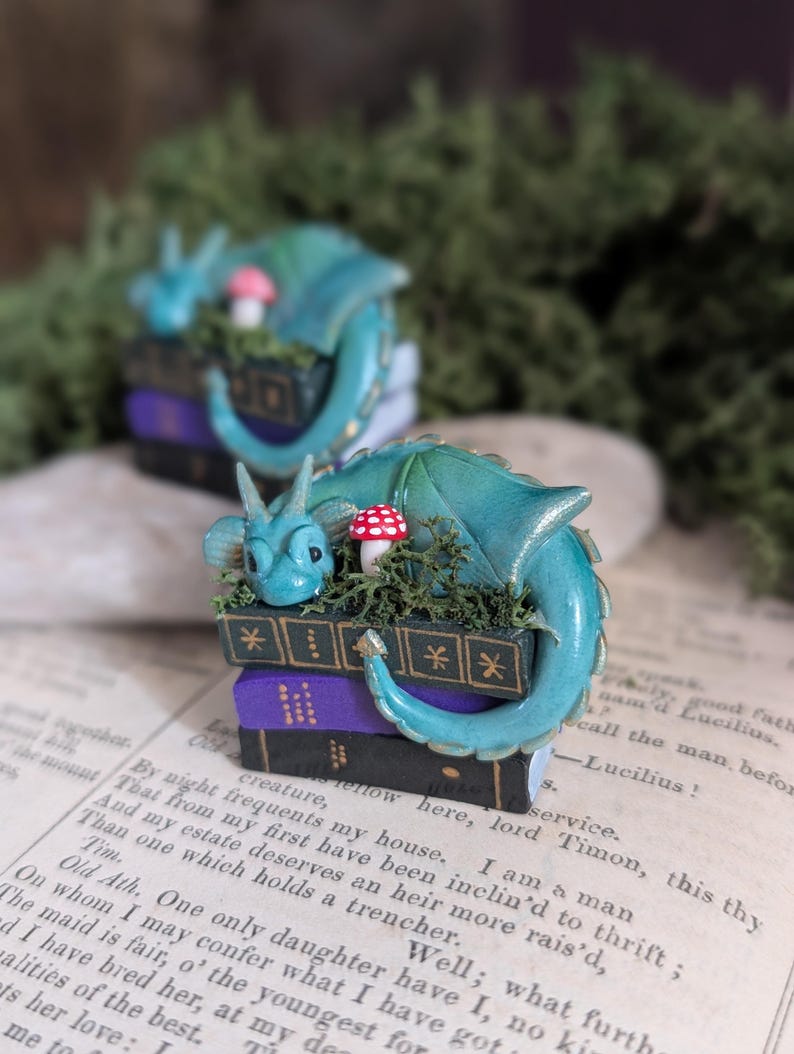 Puede incluir: Dos esculturas de fantas&iacute;a en miniatura. Cada una presenta un drag&oacute;n verde azulado enroscado alrededor de una pila de libros. Los libros son negros y morados, con un peque&ntilde;o hongo y vegetaci&oacute;n encima. El drag&oacute;n tiene una cara y una cola detalladas.