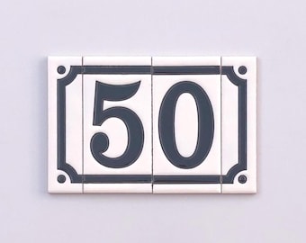 Puede incluir: Placa de número de casa de cerámica blanca con el número 50 en gris oscuro. La placa rectangular, compuesta por cuatro baldosas, tiene un borde gris oscuro y detalles decorativos en las esquinas.