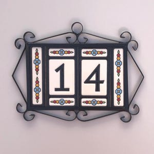 May include: Decorative house number plaque featuring the number "14" in black on white tiles. The plaque is framed in a black wrought iron design with decorative scrollwork and colorful floral accents. The overall design is rectangular with a diamond shape.
