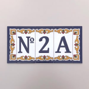 May include: A rectangular ceramic house number plaque displaying "Nº 2 A". The dark blue numerals and letters are set against a white backdrop. The plaque features a dark blue border and decorative gold and red detailing.