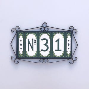 Può includere: Targa con indirizzo decorativa con il numero "31". La targa ha una cornice in ferro battuto nero con volute decorative. I numeri sono bianchi su uno sfondo di piastrelle verdi e bianche.