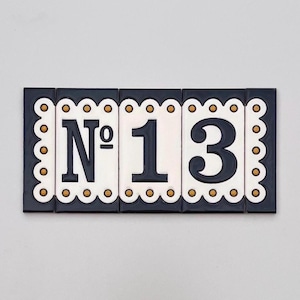 以下が含まれることがあります： 番号「13」が表示された長方形のセラミック製の家番号プレート。 プレートは3つのタイルで構成されています。 数字は白地の背景に濃い青色で、金色のアクセントが付いたスカラップの濃い青色のボーダーで囲まれています。