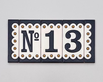 Puede incluir: Placa rectangular de cerámica con el número de casa "13". La placa está compuesta por tres baldosas. Los números son de color azul oscuro sobre un fondo blanco, enmarcados por un borde festoneado azul oscuro con detalles dorados.