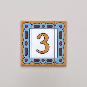 以下が含まれることがあります： 白い背景に金色の数字「3」が中央に配置された装飾タイル。 タイルは、水色のボーダー、茶色の長方形、赤い点で縁取られ、黄橙色の背景に配置されています。 タイルはおそらく番地用です。