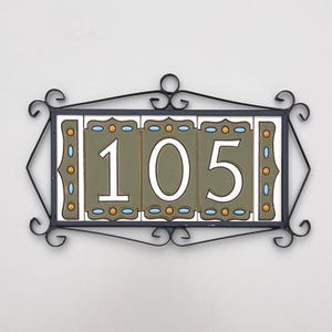 May include: Decorative address plaque featuring the number "105" in white against a green background. The plaque is framed by a black wrought iron design with decorative scrollwork and accents of orange and blue. The plaque is mounted on a white wall.