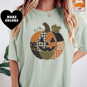 Puede incluir: Una camiseta de color verde claro con un gráfico central grande de una calabaza de Halloween. La calabaza está compuesta por varios parches estampados en naranja, negro y verde, incluyendo cuadros, lunares y estrellas. Un icono de corazón negro dice MORE COLORS.