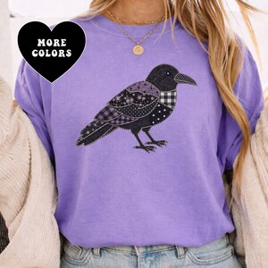 Puede incluir: Una camiseta violeta de cuello redondo con un gráfico de un cuervo compuesto por varios parches de tela en negro, blanco y morado. El diseño del ave incluye texturas de cuadros, lunares y flores. Un corazón negro con el texto MORE COLORS.