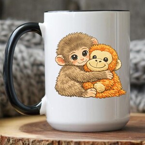 May include: White ceramic mug with a black handle and rim. The mug features a cartoon illustration of two hugging monkeys in shades of brown and orange. The mug is sitting on a wooden surface.