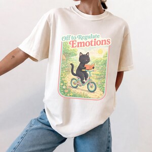 Puede incluir: Camiseta color crema con un diseño gráfico caprichoso. El diseño incluye un gato negro montando en bicicleta mientras lee un libro, sobre un fondo escénico. El texto "Off to Regulate Emotions" se muestra encima de la ilustración en una fuente juguetona.