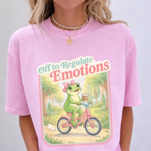 Op de afbeelding: Lichtroze t-shirt met een cartoon kikker op een roze fiets, met een koffiekopje. De kikker draagt een roze strik. Het shirt heeft de tekst "Off to Regulate Emotions" in retro stijl.