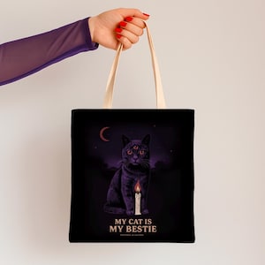 Puede incluir: Bolso tote negro con una ilustración de un gato de tres ojos, una vela y el texto "MY CAT IS MY BESTIE". El gato está sobre un fondo de cielo nocturno púrpura oscuro con una luna creciente. El bolso tiene asas color crema.