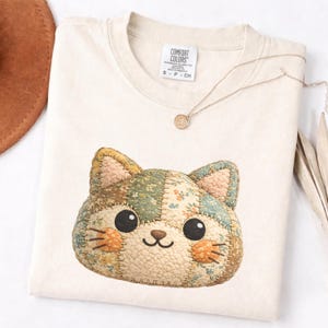 Puede incluir: Camiseta color crema con un diseño de cara de gato patchwork. La cara del gato presenta una mezcla de telas florales y estampadas en tonos verdes, marrones y melocotón. La camiseta tiene cuello redondo y la marca "Comfort Colors" es visible.