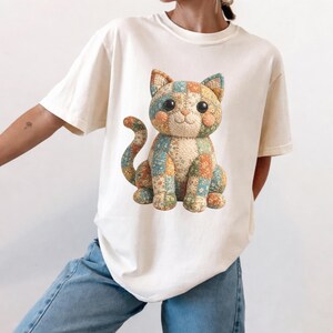 Puede incluir: Camiseta color crema con un diseño de gato patchwork colorido. El gato está sentado, con una cara sonriente y una larga cola. El diseño de patchwork incluye cuadrados naranjas, azules y amarillos. La camiseta es de estilo casual y cómodo.