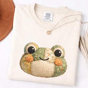 Può includere: T-shirt color crema con un design a forma di faccia di rana patchwork. La rana ha grandi occhi neri e il patchwork include motivi floreali verdi, gialli e arancioni. Una collana d'oro con un piccolo ciondolo è appoggiata sulla maglietta.