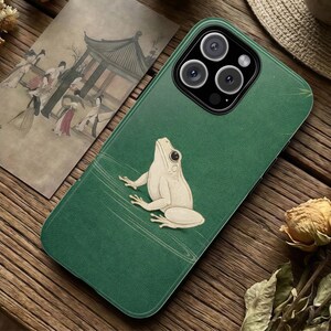 Puede incluir: Funda de teléfono verde con un diseño de rana crema. La funda tiene un aspecto texturizado y una ilustración de rana en relieve. La funda del teléfono está sobre una superficie de madera con una ilustración de estilo vintage y flores secas.