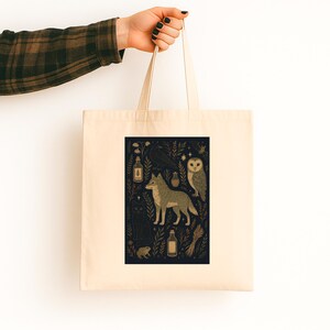 May include: Cream-colored tote bag featuring a dark blue graphic with gold and cream illustrations. The design includes a wolf, owl, raven, cat, and bottles. The bag is held by a person with a plaid sleeve and black nail polish.