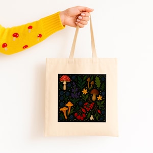 May include: A beige tote bag with a square, black graphic featuring colorful mushrooms, berries, and foliage. The mushrooms are red, orange, and brown. The bag is held by a person wearing a yellow sweater with red mushroom designs.