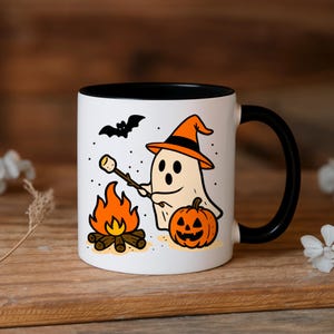 Puede incluir: Taza de cerámica blanca con interior y asa negros, con una ilustración de Halloween. El diseño incluye un fantasma asando una malvavisco sobre una fogata, una calabaza y un murciélago. La taza está sobre una superficie de madera.