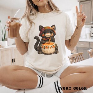 Puede incluir: Camiseta de color marfil con una ilustración caprichosa de un gato negro con un pañuelo y removiendo sopa en un cuenco decorativo. El gato es el foco central, con una cuchara de madera. La camiseta es de corte holgado.