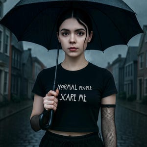 Puede incluir: Una persona con una camiseta negra con el texto blanco "NORMAL PEOPLE SCARE ME", sosteniendo un paraguas negro. El fondo muestra una calle lluviosa con edificios.