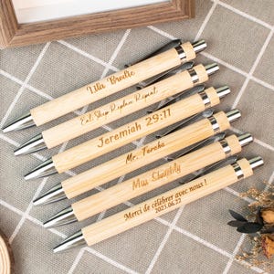 May include: A collection of engraved bamboo pens with silver accents. Each pen features unique text, including names and phrases like "Eat Sleep Replace Repeat" and "Jeremiah 29:11". The pens are arranged on a checkered surface.