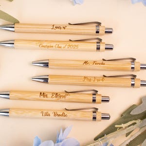May include: Six bamboo pens with silver accents are arranged on a light beige surface, surrounded by blue floral decorations. Each pen is engraved with a name or message, including "Lauren", "Christopher Class of 2025", and "Mr. Teroha".