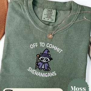 May include: A moss green t-shirt with the embroidered phrase "OFF TO COMMIT SHENANIGANS" and a cartoon raccoon wearing a wizard hat. The shirt also has the word "EMBROIDERED" and the color "Moss" displayed.