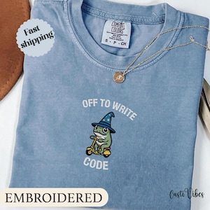 May include: A light blue t-shirt with the embroidered phrase "OFF TO WRITE CODE" and a cartoon frog wearing a wizard hat riding a scooter. A gold necklace with a pendant is draped across the shirt. The shirt has a Comfort Colors tag.