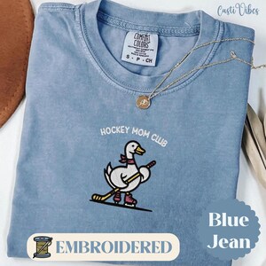 Könnte beinhalten: Blaues Jeans-T-Shirt mit der Aufschrift "HOCKEY MOM CLUB" über einer gestickten Cartoon-Ente, die Schlittschuhe trägt und einen Hockeyschläger hält. Das Shirt hat ein Comfort Colors-Etikett und das Wort "EMBROIDERT" steht unten.