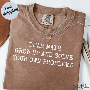 May include: A brown t-shirt with the slogan "DEAR MATH GROW UP AND SOLVE YOUR OWN PROBLEMS" in white. A silver necklace with a pendant rests on the shirt. The t-shirt has a Comfort Colors label.