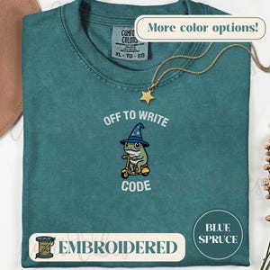 May include: A blue spruce colored t-shirt featuring an embroidered graphic of a green frog wearing a blue wizard hat and riding a yellow scooter. The text above the frog reads OFF TO and below reads WRITE CODE. The shirt has a Comfort Colors label.