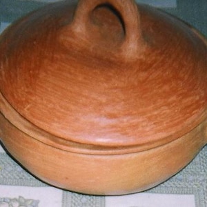 Può includere: Una pentola da cucina in terracotta fatta a mano con coperchio. La pentola è di un caldo colore marrone terroso e presenta due manici sui lati e uno sul coperchio. La pentola viene probabilmente utilizzata per cucinare e servire cibo.