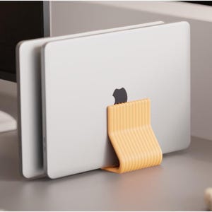 May include: A silver laptop is held upright by a yellow-orange stand. The stand has a curved, ribbed design. The Apple logo is visible on the laptop. The setup is on a gray surface, with a computer monitor in the background.