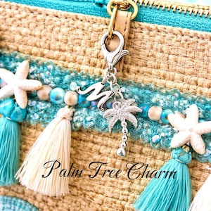 May include: Close-up of a woven bag with a turquoise zipper and decorative trim. The trim includes white starfish, turquoise tassels, and a silver palm tree charm. The text "Palm Tree Charm" is visible.